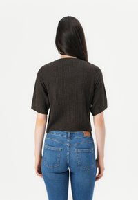 Kvinna med långt mörkt hår som bär en ribbad kortärmad mörkbrun topp och blå högmidjade jeans, vänd bort från kameran mot en vit bakgrund.