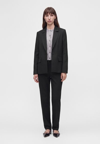 Femme debout, face à l’avant, portant un costume noir avec un pantalon sur mesure, un blazer à un bouton, une chemise grise boutonnée et des ballerines noires pointues.
