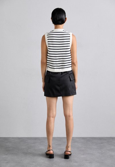 Άτομο με κοντά μαύρα μαλλιά, που φοράει ένα αμάνικο λευκό μπλουζάκι με μαύρες ρίγες, μια μαύρη μίνι φούστα και μαύρες γόβες, στέκεται σε γκρι δάπεδο.