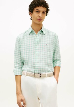 Uomo che indossa una camicia a quadri bianca e verde chiaro con le maniche arrotolate e pantaloni bianchi con cintura intrecciata beige, in piedi davanti a uno sfondo semplice.