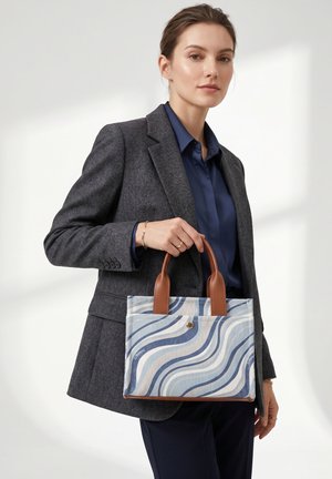 Femme portant un blazer gris et une chemise bleu marine tient un sac à main bleu et blanc à motifs avec des poignées marron sur un fond uni.