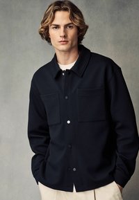 Schwarze strukturierte Button-Up-Jacke mit zwei vorderen Taschen und einem Kragen. Der Stoff hat ein gepolstertes Aussehen und ist mit einem weißen Hemd darunter kombiniert.