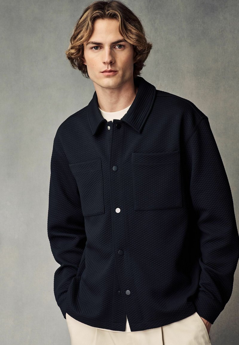 Schwarze strukturierte Button-Up-Jacke mit zwei vorderen Taschen und einem Kragen. Der Stoff hat ein gepolstertes Aussehen und ist mit einem weißen Hemd darunter kombiniert.