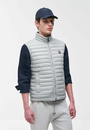 Jonge man die een lichtgrijze doorgestikte bodywarmer draagt over een marineblauw overhemd, lichtgrijze broek en een marineblauw honkbalpetje, staand tegen een effen achtergrond.