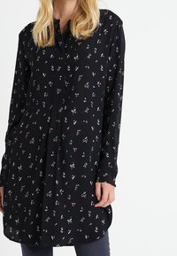 Svart blus med blommönster i rosa och vitt. Den har en knappstängning fram, långa ärmar och en rundad nederkant. Tillverkad av lätt tyg.