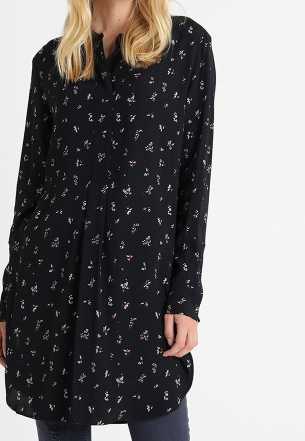 Svart blus med blommönster i rosa och vitt. Den har en knappstängning fram, långa ärmar och en rundad nederkant. Tillverkad av lätt tyg.