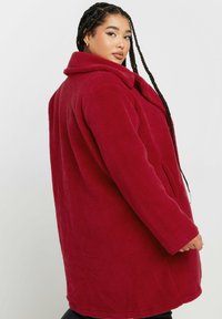 Cappotto in pile rosso con colletto, maniche lunghe e tasche laterali. Caratterizzato da una texture morbida e una vestibilità ampia, arriva a lunghezza metà coscia.
