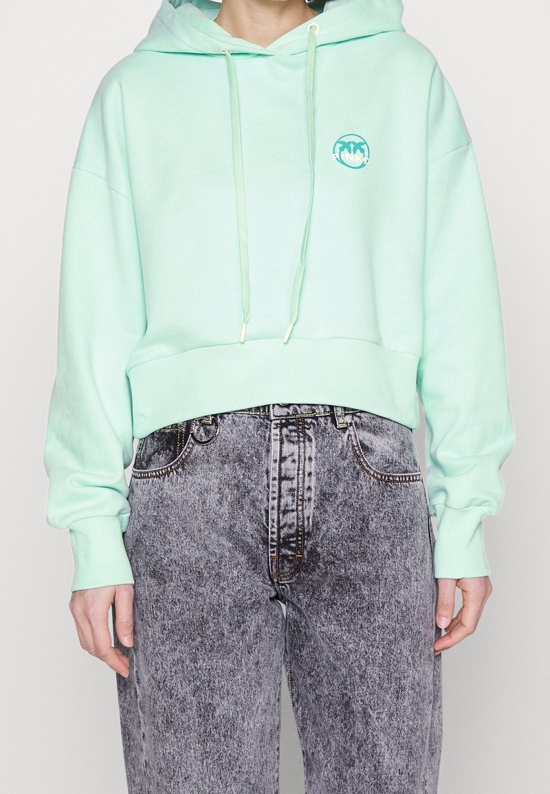 Miętowa bluza z kapturem o kroju cropped, z prążkowanymi mankietami i regulowanym kapturem. Ma okrągłe logo na klatce piersiowej oraz noszone czarne dżinsy.
