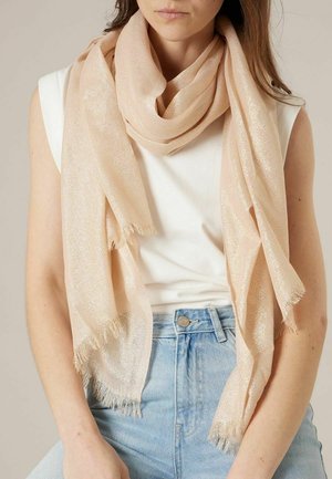 Femme portant une écharpe scintillante beige clair drapée autour du cou sur un haut blanc sans manches et un jean bleu clair.