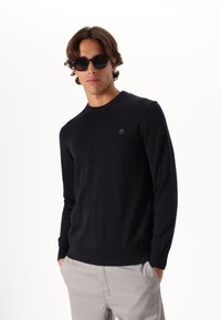 Maglione girocollo blu scuro in tessuto morbido con polsini e orlo a coste, dotato di un piccolo logo sul petto. Abbinato a pantaloni grigi chiari.