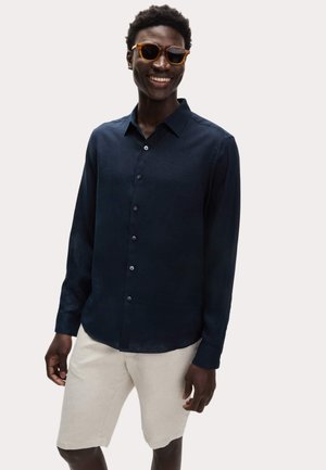 Uśmiechnięty mężczyzna w pomarańczowych okularach przeciwsłonecznych, ciemnoniebieskiej koszuli na guziki i jasnobeżowych szortach na jednolitym jasnym tle.