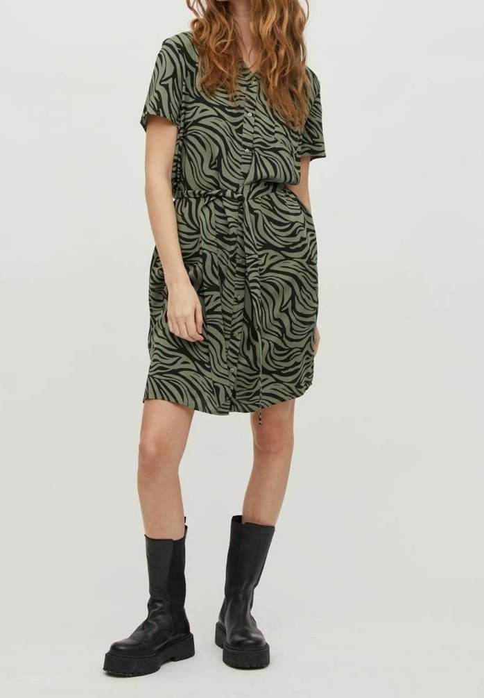 Femme portant une robe à manches courtes, boutonnée, à imprimé animalier avec un lien à la taille et des bottes noires épaisses mi-mollet, debout devant un fond uni.