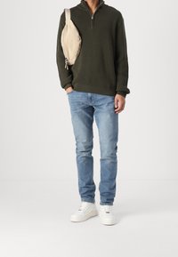 Pull-over en maille vert olive avec col zippé, jean slim bleu clair et baskets blanches. Sac bandoulière beige avec détail zippé.