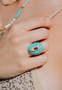 Bague en turquoise avec un accent rouge et un sertissage en or. La main repose contre un top beige perlé. Texture lisse avec de fins détails.