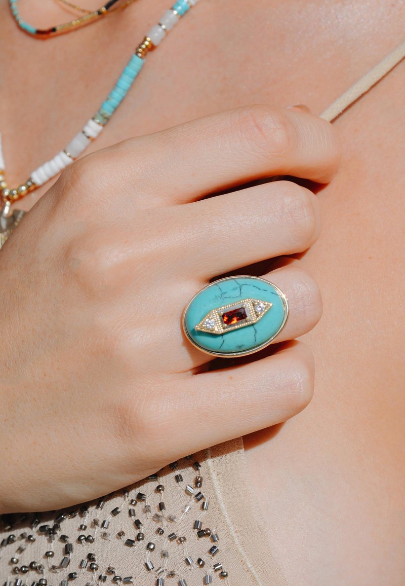 Bague en turquoise avec un accent rouge et un sertissage en or. La main repose contre un top beige perlé. Texture lisse avec de fins détails.