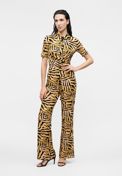 Γυναίκα ποζάρει με ολόσωμη φόρμα με κοντά μανίκια και γιακά, με έντονο κίτρινο, μαύρο και μπεζ αφηρημένο ριγωτό σχέδιο και μαύρα πέδιλα με ψηλό τακούνι.