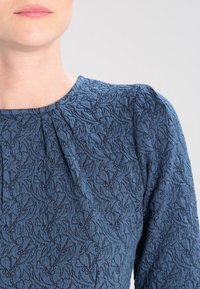 Haut bleu marine texturé avec une broderie florale complexe, des fronces au niveau de l'encolure et des manches légèrement bouffantes.