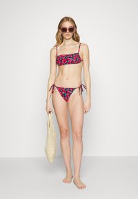 Conjunto de bikini floral com parte de cima bandeau e parte de baixo com laços laterais, apresentando um fundo preto, flores em tons de rosa e azul, e uma bolsa de palha.