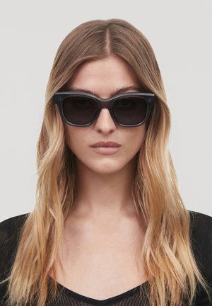 Black rectangular sunglasses with thick frames and dark lenses, set against a neutral background, worn by a model with long hair.