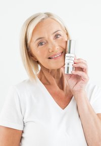 Klar glasflaska med silverlock märkt "âme pure Collagen Therapy." Modellen bär en vit T-shirt med V-ringning och ler.