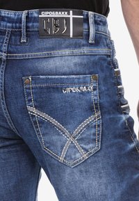 Dunkelblaue Jeans aus Denim mit einem verwaschenen Finish. Sie verfügen über ein Lederpatch an der Taille, dekorative Nähte an den Gesäßtaschen und das Markenlogo.
