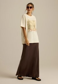 Kremowa, oversize'owa koszulka z graficznym motywem palmy oraz napisem "Paradis", zestawiona z długą brązową spódnicą i czarnymi sandałami.