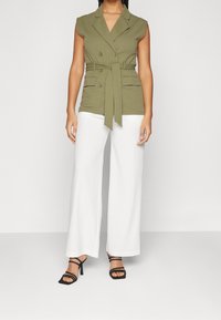 Zeleno-olivový bezrukávový blazér s dvojitými knoflíky, páskem v pase a dvěma předními kapsami; kombinovaný s širokými bílými kalhotami a černými sandály.