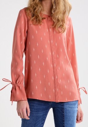 Femme portant un chemisier rose saumon à manches longues avec de petits motifs blancs et des poignets noués, associé à un jean bleu sur un fond uni.