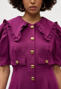 Vestido roxo com um grande colarinho com bordas franzidas, mangas curtas bufantes e detalhes de botões em tom de ouro na frente e nos bolsos.
