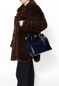 Borsa finta in pelle di coccodrillo di colore blu navy, con dettagli in metallo dorato, forma strutturata e doppi manici, abbinata a un cappotto marrone e pantaloni neri.