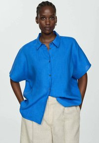 Camicia azzurra a maniche corte con chiusura a bottoni, vestibilità rilassata e colletto, realizzata in tessuto leggero. Abbinata a pantaloni beige.