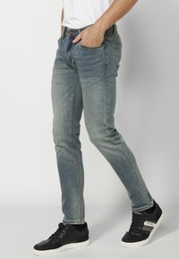 Mršavi traperice u izblijedjelom plavom denimu, sa pet džepova, petljama za remen i zatvaranjem na zatvarač. Uparene s crnim tenisicama.