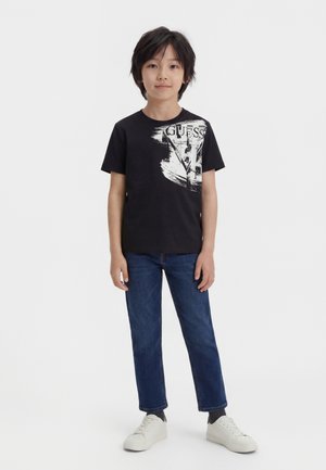 Fekete grafikás pólót, kék farmert és fehér tornacipőt viselő fiú áll tiszta fehér háttér előtt.