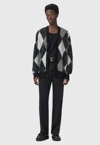 Cardigan en tricot fluffy à motif argyle noir et gris, avec un devant ouvert, des boutons noirs et des poignets côtelés, porté par-dessus un haut et un pantalon noirs.