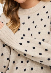 Pull beige en tricot avec des pois noirs duveteux porté par une personne aux longs cheveux châtain clair, montrant une partie du visage et les bras croisés.