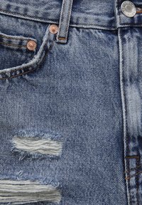 Gros plan sur un jean en denim bleu avec une petite poche avant, une boucle de ceinture, des rivets métalliques et deux déchirures horizontales usées montrant des fils blancs.