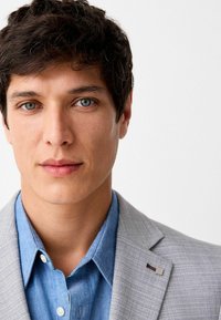 Hellgrauer Blazer über einem blauen Denim-Hemd. Der Blazer hat eine feine Textur; das Hemd hat einen klassischen Kragen und Knöpfe. Glatte Haut und neutraler Ausdruck.