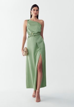 Frau in grünem Satin-Einschultrigem Kleid mit hohem Schlitz, die eine geflochtene Clutch hält, goldene Ohrringe trägt und Riemchen-Sandaletten mit hohen Absätzen anhat.