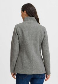 Veste en mélange de laine grise avec un col haut, une silhouette ajustée et des coutures structurées, portée sur un jean bleu, vue de dos.