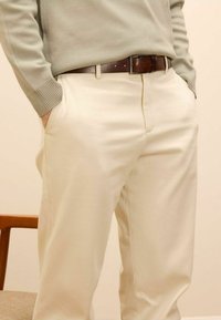 Lichtbeige broek met een gladde textuur, gecombineerd met een groene gebreide trui. Een bruine leren riem met een klassieke gesp maakt de look compleet.