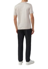 Man die met zijn rug naar de kijker staat, gekleed in een lichtbeige overhemd met korte mouwen, zwarte broek en witte sneakers, op een witte achtergrond.