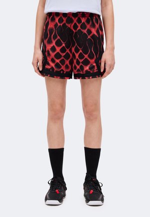 Person iført røde og sorte mønstrede shorts, sorte sokker, sorte sneakers og en hvid top, stående mod en ensfarvet lys baggrund.