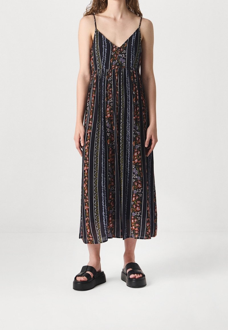 Robe maxi à fines bretelles, fond marine, rayures verticales, motifs floraux rouges et verts, coupe ample, associée à des sandales à plateforme noires.