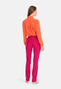 Long-sleeve, collarless orange blouse paired with high-waisted, textured pink trousers. Footwear is beige with a short, slender heel.
