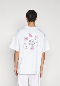 Tricou alb din bumbac, prezentând o ilustrație colorată cu fluturi, flori și mâini pe spate, cu o croială lejeră și mâneci scurte.