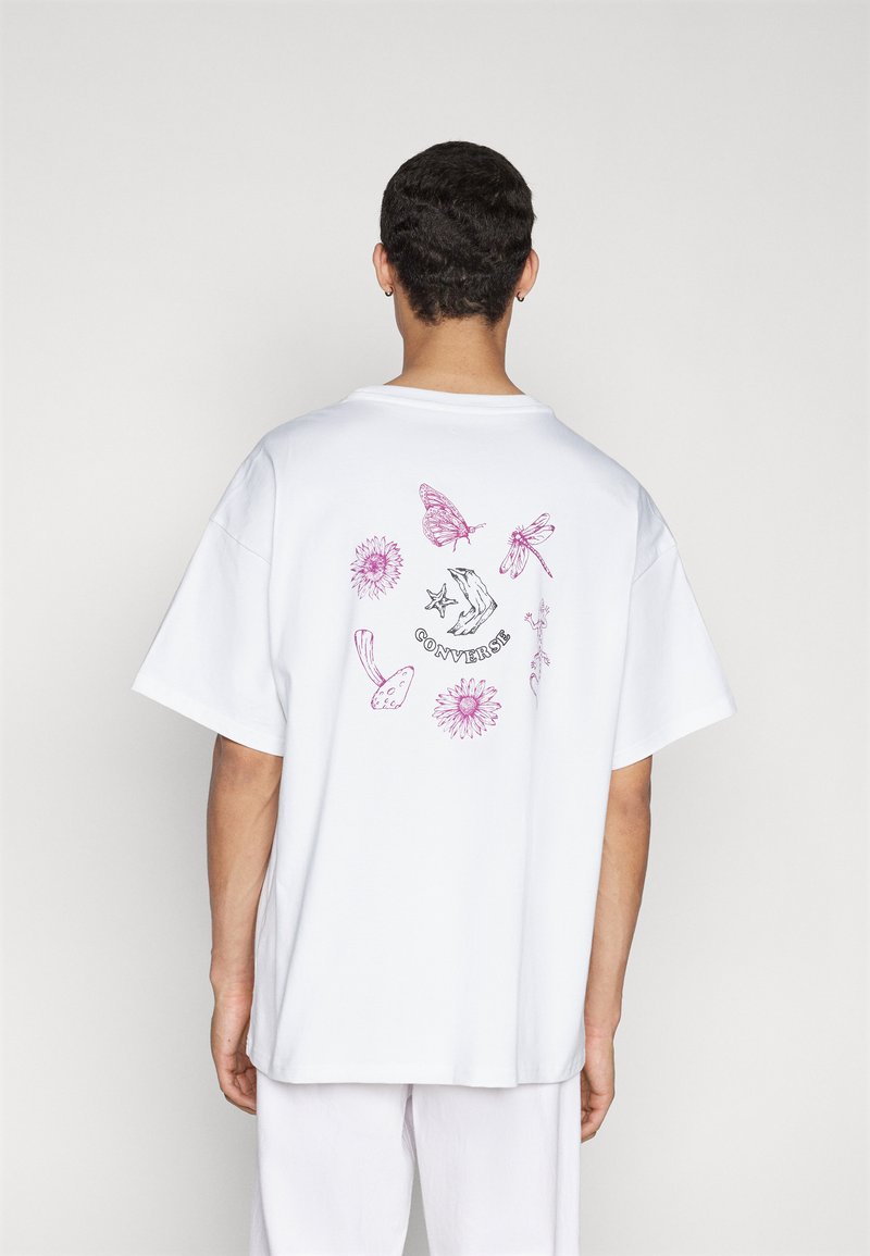 Tricou alb din bumbac, prezentând o ilustrație colorată cu fluturi, flori și mâini pe spate, cu o croială lejeră și mâneci scurte.