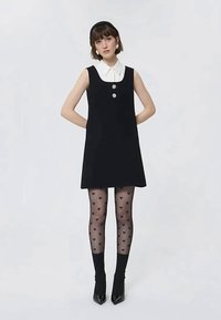 Femme portant une robe sans manches noire avec un col blanc, des boutons floraux, des collants à motifs de cœurs, des chaussettes noires à la cheville et des talons noirs pointus.