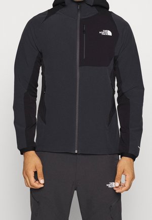 Homme portant une veste à capuche zippée noire et gris foncé et un pantalon assorti avec les logos The North Face sur la poitrine et la cuisse.