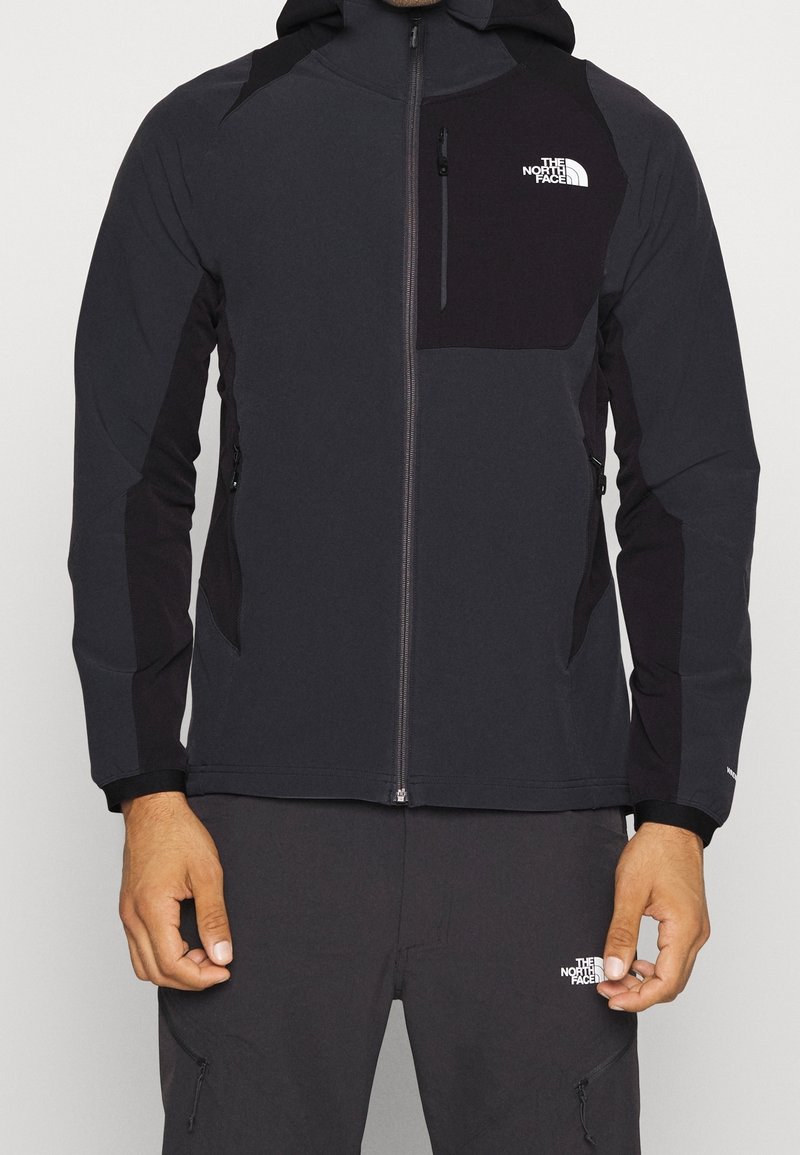 Homme portant une veste à capuche zippée noire et gris foncé et un pantalon assorti avec les logos The North Face sur la poitrine et la cuisse.