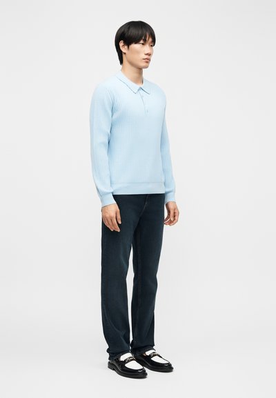 Jeune homme debout portant un polo à manches longues côtelé bleu clair, un jean foncé et des mocassins en cuir noir et blanc.